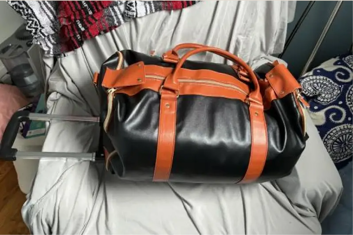 A black and tan Luhxe bag with its wheels attached, resting on a bed